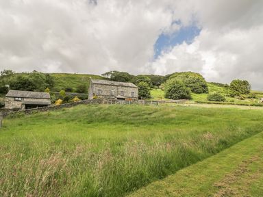 A grassy field with stone buildings and trees on a hill at Bank End Barn in Grizebeck