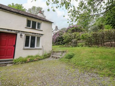 A white house with a red door next to a gravel driveway and grassy garden at Hall Bank Cottage in Rydal near Ambleside
