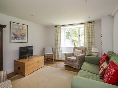 A living room with a green sofa with cushions a striped armchair a wooden chest used as a tv stand a wicker chair and curtains at Hall Bank Cottage in Rydal near Ambleside