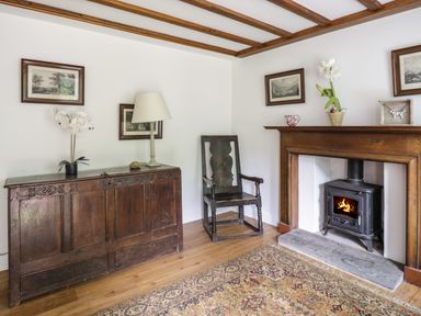 A corner of a room with a wooden chest, a lamp, a wooden chair, a fireplace with a wood stove, and framed pictures on the walls at Hall Bank Cottage in Rydal near Ambleside