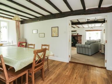 A dining room with a table covered by a green polka dot cloth and wooden chairs open to a living room with sofas and a fireplace at Hall Bank Cottage in Rydal near Ambleside