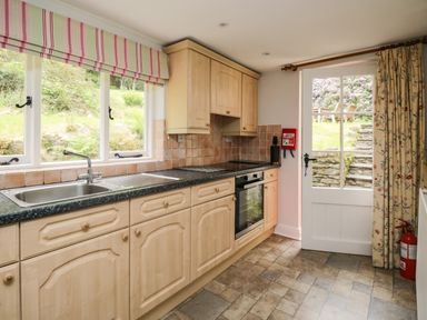 A kitchen with wooden cabinets a stainless steel sink an oven a door leading outside and patterned curtains at Hall Bank Cottage in Rydal near Ambleside