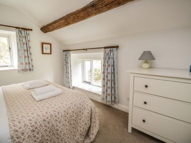 A bedroom with a bed covered in a floral quilt next to windows with floral curtains and a white dresser with a lamp at Hall Bank Cottage in Rydal near Ambleside