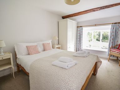 A bedroom with a double bed white and pink bedding bedside tables lamps a white wardrobe and a window with blue floral curtains at Hall Bank Cottage in Rydal near Ambleside