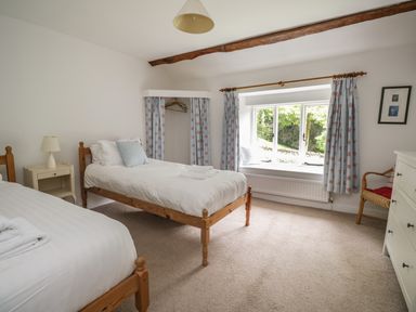 A bedroom with two single beds a window with floral curtains and a wicker chair at Hall Bank Cottage in Rydal near Ambleside