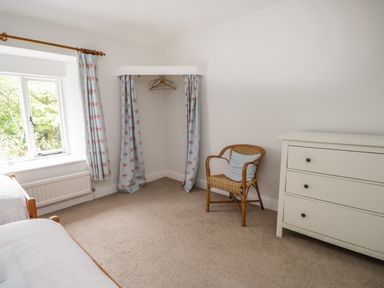 A bedroom with a window with curtains a wicker chair with a cushion a white dresser and a corner closet with curtains at Hall Bank Cottage Rydal near Ambleside