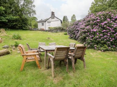 A wooden outdoor table with six chairs on grass near a flowering bush and a white stone house at Hall Bank Cottage in Rydal near Ambleside
