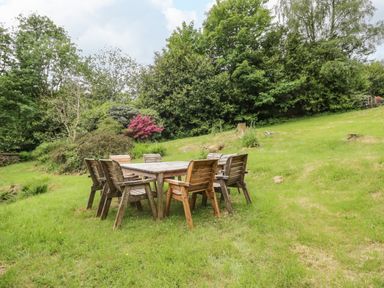 An outdoor wooden table with eight wooden chairs on grass surrounded by trees and shrubs at Hall Bank Cottage in Rydal near Ambleside