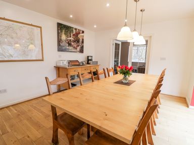 A dining room with a long wooden table and chairs a vase with flowers on the table a sideboard with toasters coffee maker and bread box two paintings on the wall and ceiling lights at Ty Coch in Llanberis