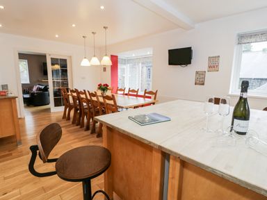 A dining room with a long wooden table and chairs a kitchen island with stools and a bottle of prosecco at Ty Coch in Llanberis