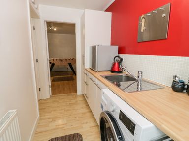 A narrow kitchen with a washing machine sink kettle and fridge looking into a room with a foosball table at Ty Coch in Llanberis