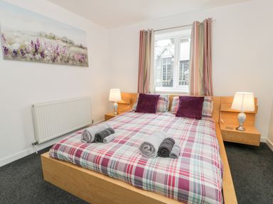 A bedroom with a wooden bed with plaid bedding and rolled towels on it two bedside tables with lamps and a window with striped curtains at Ty Coch in Llanberis