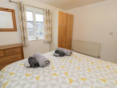 A bedroom with a bed covered in a patterned bedspread and rolled towels a wooden dresser a wardrobe a window with patterned curtains and a radiator on the wall at Ty Coch in Llanberis