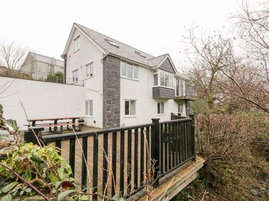 A white multi-story house with a wooden deck and black railing surrounded by trees and shrubs at Ty Coch in Llanberis