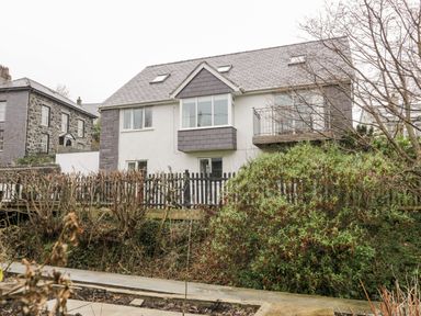A two-story house with gray roof and white walls behind a wooden fence and bushes at Ty Coch in Llanberis