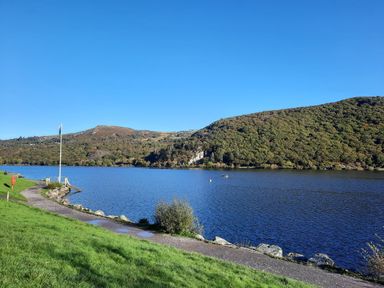 A path alongside a lake with a flagpole and hills in the background at Ty Coch in Llanberis