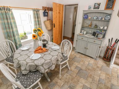 A dining area with a round table set with plates, croissants, and a coffee press near a window with plaid curtains and a gray cabinet with decorative items in Snowdrop Cottage in Laugharne