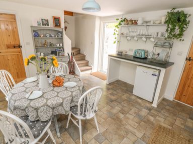 A dining area with a round table set with plates and croissants a hutch with dishes stairs and a countertop with a toaster and mini fridge at Snowdrop Cottage in Laugharne