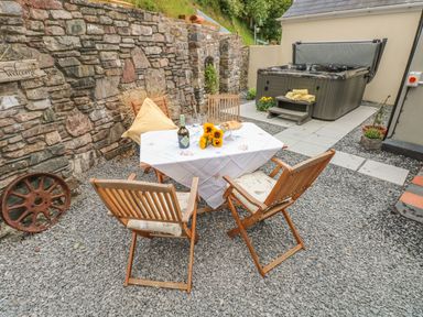 An outdoor patio area with a table and four wooden chairs a hot tub and stone wall at Snowdrop Cottage in Laugharne