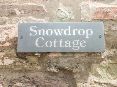 A stone wall with a grey rectangular sign reading Snowdrop Cottage in Laugharne