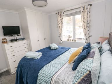 A bedroom with a bed towels on a blue blanket a white dresser a wall-mounted TV and a window with patterned curtains at The Retreat Wisemans Bridge near Saundersfoot