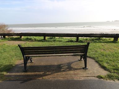 A wooden bench on a paved area facing the sea with a grass area and wooden rail in the background at The Retreat in Wisemans Bridge near Saundersfoot