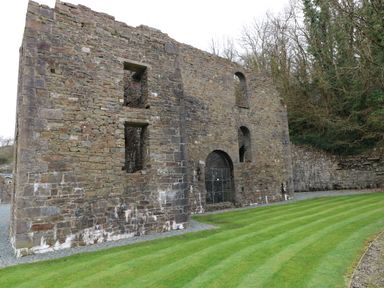 An old stone building with window openings and an arched gated entrance on a grass lawn next to trees at The Retreat in Wisemans Bridge near Saundersfoot
