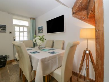 A dining area with six chairs around a table with a white tablecloth and a flower centerpiece wooden tripod lamp and wall mounted television at Curlews Cottage in Looe