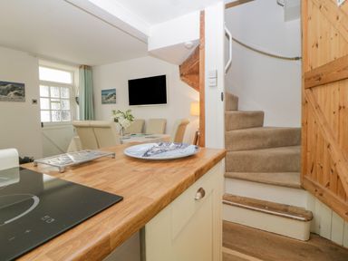 A kitchen counter with a stove and a plate overlooking a dining area with chairs a television and a staircase at Curlews Cottage in Looe