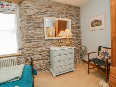 A bedroom corner with a white chest of drawers a mirror above a stone wall a chair with a pillow and a bed with blankets at Curlews Cottage in Looe