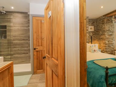 A bathroom with a shower and wooden doors next to a bedroom with stone walls and a bed with a teal blanket at Curlews Cottage in Looe
