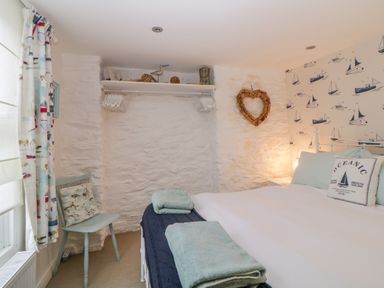 A bedroom with a bed with pillows and folded towels a chair with a cushion curtains a heart-shaped wall decoration and boat patterned wallpaper at Curlews Cottage in Looe