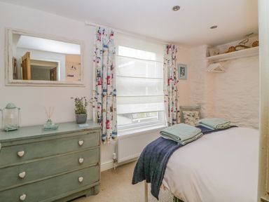 A bedroom with a bed with folded towels a chest of drawers a window with patterned curtains and a wall shelf at Curlews Cottage in Looe