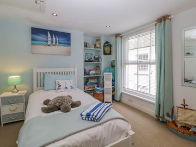 A bedroom with a single bed, bedside table with lamp, shelves with books and toys, large window with curtains and a toy ship on the floor at Curlews Cottage in Looe