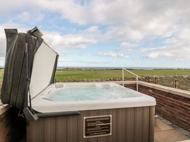 An outdoor hot tub with cover partially open overlooking a grassy field and ocean at Bank Top Cottage in Embleton