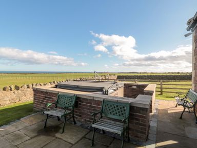 An outdoor patio with a hot tub surrounded by brick walls and three green metal benches at Bank Top Cottage in Embleton