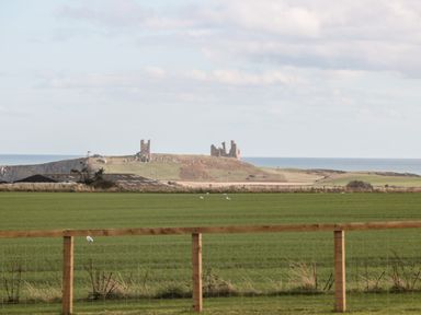 A green field with a wooden fence and distant castle ruins on a hill by the sea at Bank Top Cottage in Embleton