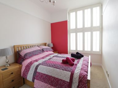 A bedroom with a wooden bed frame pink and purple bedding bedside tables with lamps and large windows with white shutters at 1 Beachtop Court Tenby