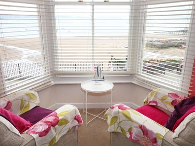 A seating area with two armchairs a small round table and windows with blinds overlooking a beach at 1 Beachtop Court in Tenby