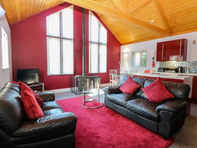 A living room with black leather sofas and red cushions near a glass coffee table and a TV with a red carpet and wooden ceiling at Rookyard Cottage in Holton near Halesworth
