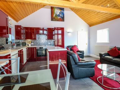 An open-plan kitchen and living area with red cabinets black leather sofas and a glass dining table at Rookyard Cottage in Holton near Halesworth