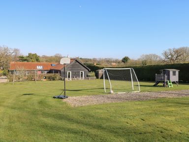 A backyard with a basketball hoop, soccer goal, swing set, playhouse, and a house with a red roof at Rookyard Cottage in Holton near Halesworth