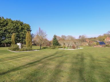 A garden with trimmed grass shrubs trees and a trellis structure at Rookyard Cottage in Holton near Halesworth