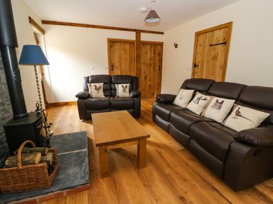 A living room with brown leather sofas with animal print cushions a wooden coffee table a wood stove with a basket of firewood and wooden doors at Ploony Hill Lodge in Bleddfa