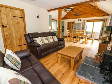 A living room with brown leather sofas, wooden coffee table, dining table with chairs, wood stove, and kitchen area at Ploony Hill Lodge in Bleddfa