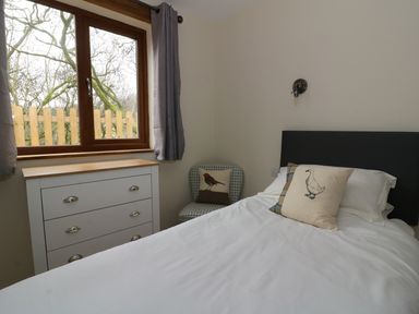 A bedroom with a bed with white bedding and animal print pillows a chest of drawers a patterned chair and a window with gray curtains at Ploony Hill Lodge in Bleddfa