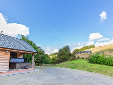 An outdoor area with a wooden shelter housing a hot tub a gate and trees near a grassy hillside at Ploony Hill Lodge in Bleddfa