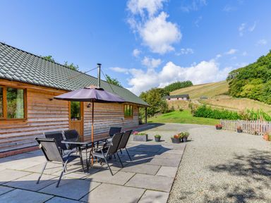 An outdoor patio with a table and chairs under an umbrella next to a wooden building at Ploony Hill Lodge in Bleddfa