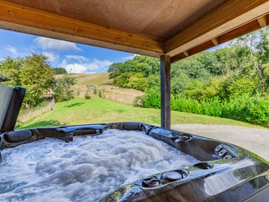 A bubbling hot tub on a covered patio overlooking a grassy yard and trees at Ploony Hill Lodge in Bleddfa