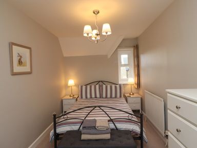 A bedroom with a bed flanked by two bedside tables with lamps a window a chest of drawers and a framed picture at Jasmine Cottage Lesbury near Alnmouth